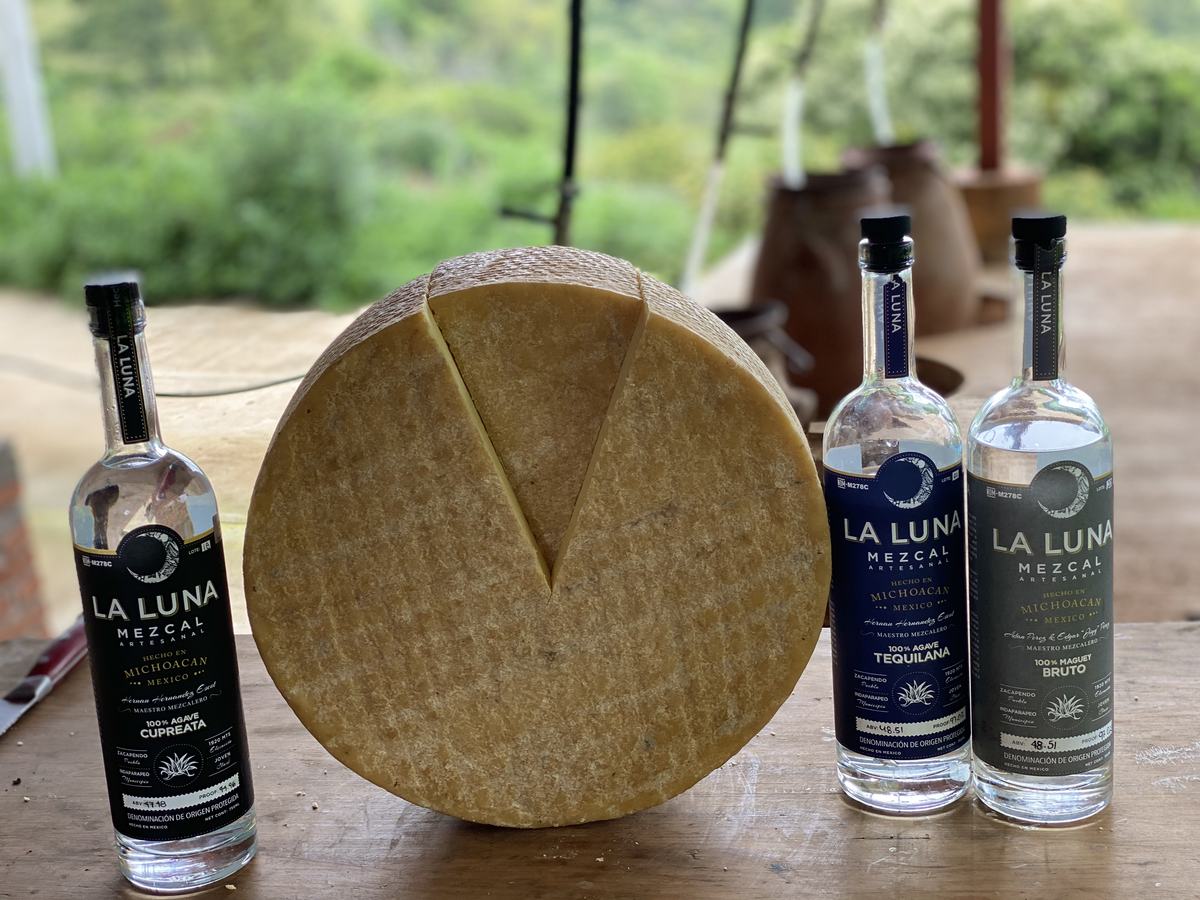 Three bottles of La Luna Mezcal beside a cut wheel of aged cheese on a wooden table at the vinata, green hills behind.