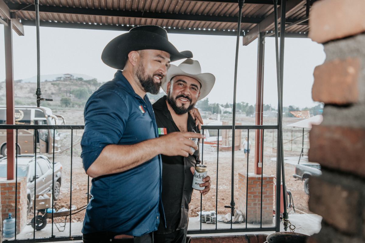 Salvador and Adrian, arm in arm and laughing, on a covered terrace above dry hills.