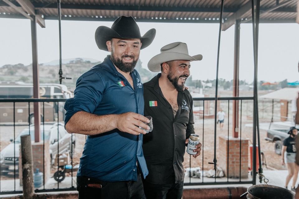 Salvador and Adrian side by side and smiling, one raising a small cup, on the same terrace.