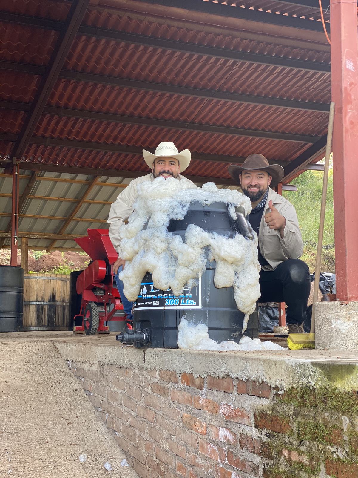 Salvador Picazo Chavez and Adrian Gallegos crouch behind a fermentation drum as thick foam spills over its rim, under the tin roof of the vinata.
