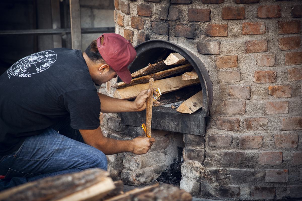 A producer kneels at the brick firebox beneath a still, feeding split pine into the fire, a stack of cut wood in the foreground.