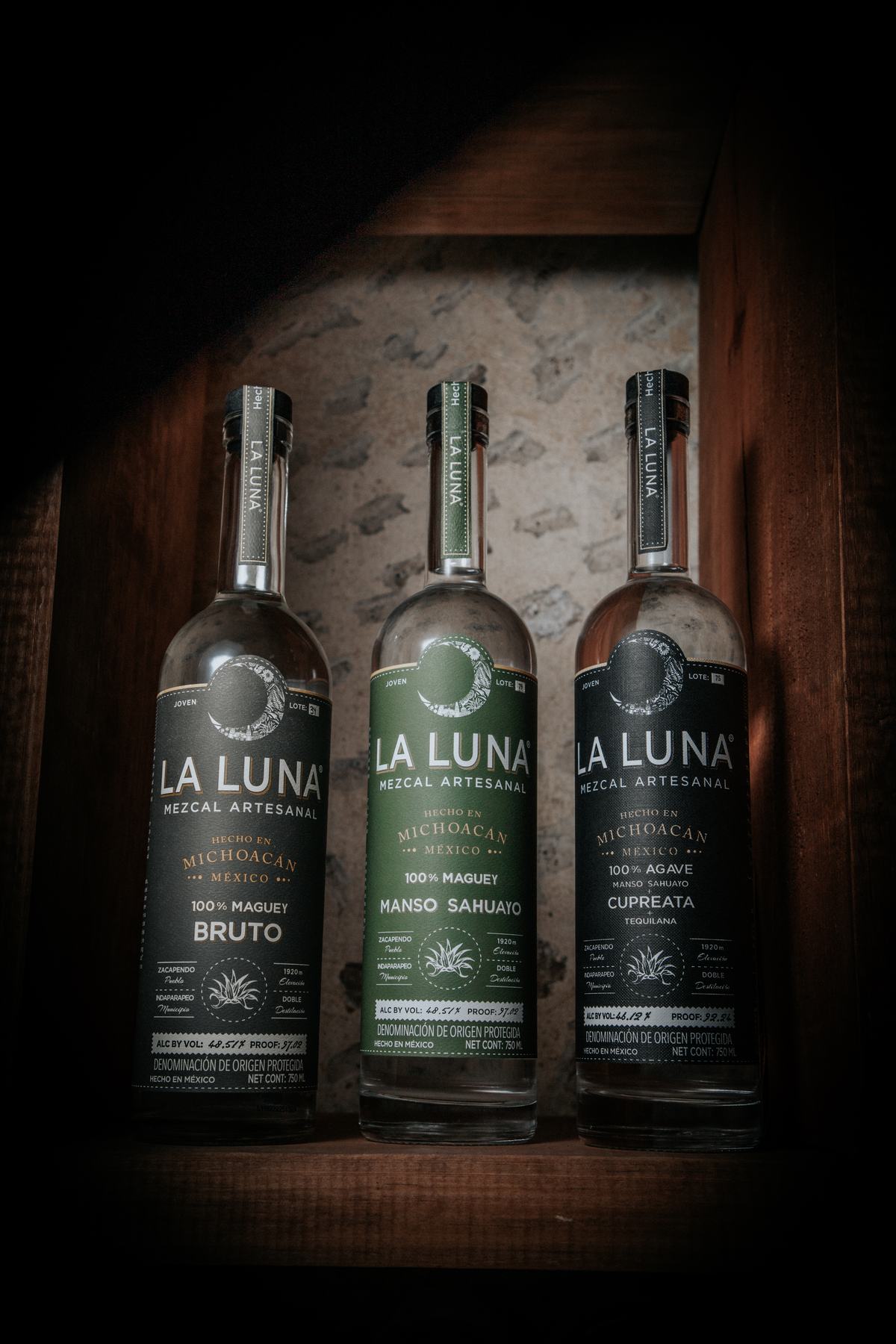 Three bottles of La Luna Mezcal — Bruto, Manso Sahuayo and Cupreata — standing on a dark wooden shelf against a weathered stone wall.