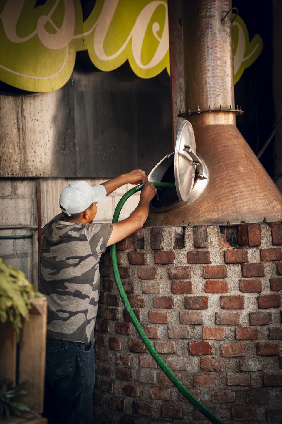 A producer reaches up to feed a hose into the open hatch of a copper still set in brick.