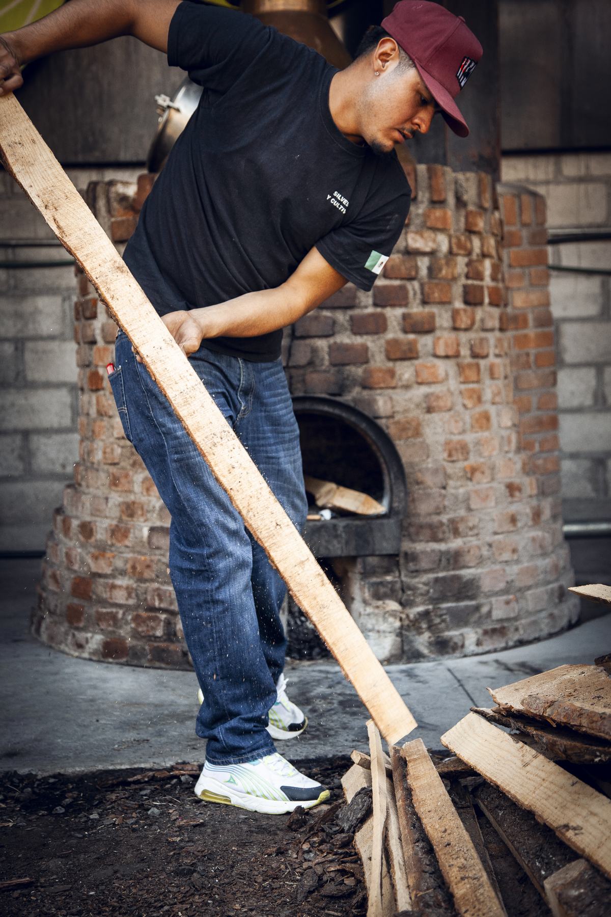A producer hauls a long plank of firewood past the brick firebox of a copper still.
