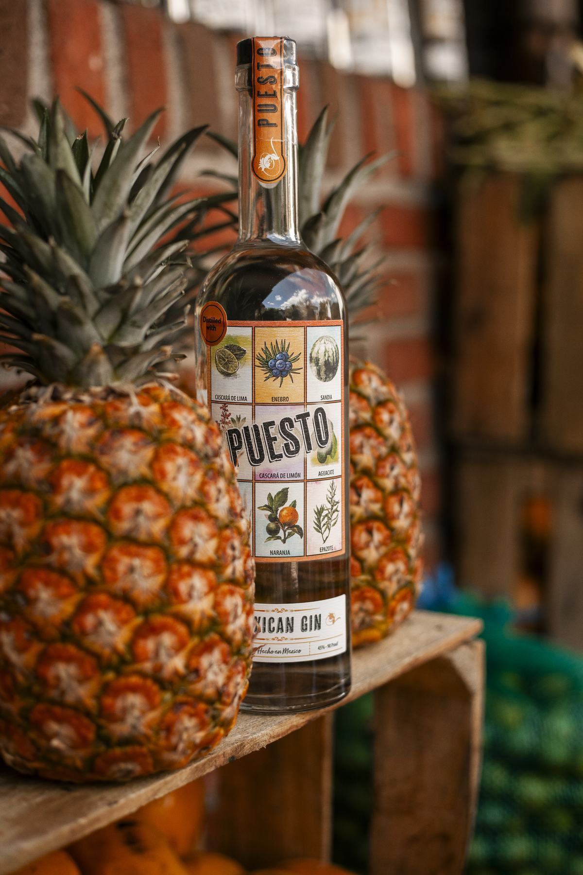 A bottle of Puesto Mexican Gin on a wooden crate beside whole pineapples at a brick-walled fruit stand.