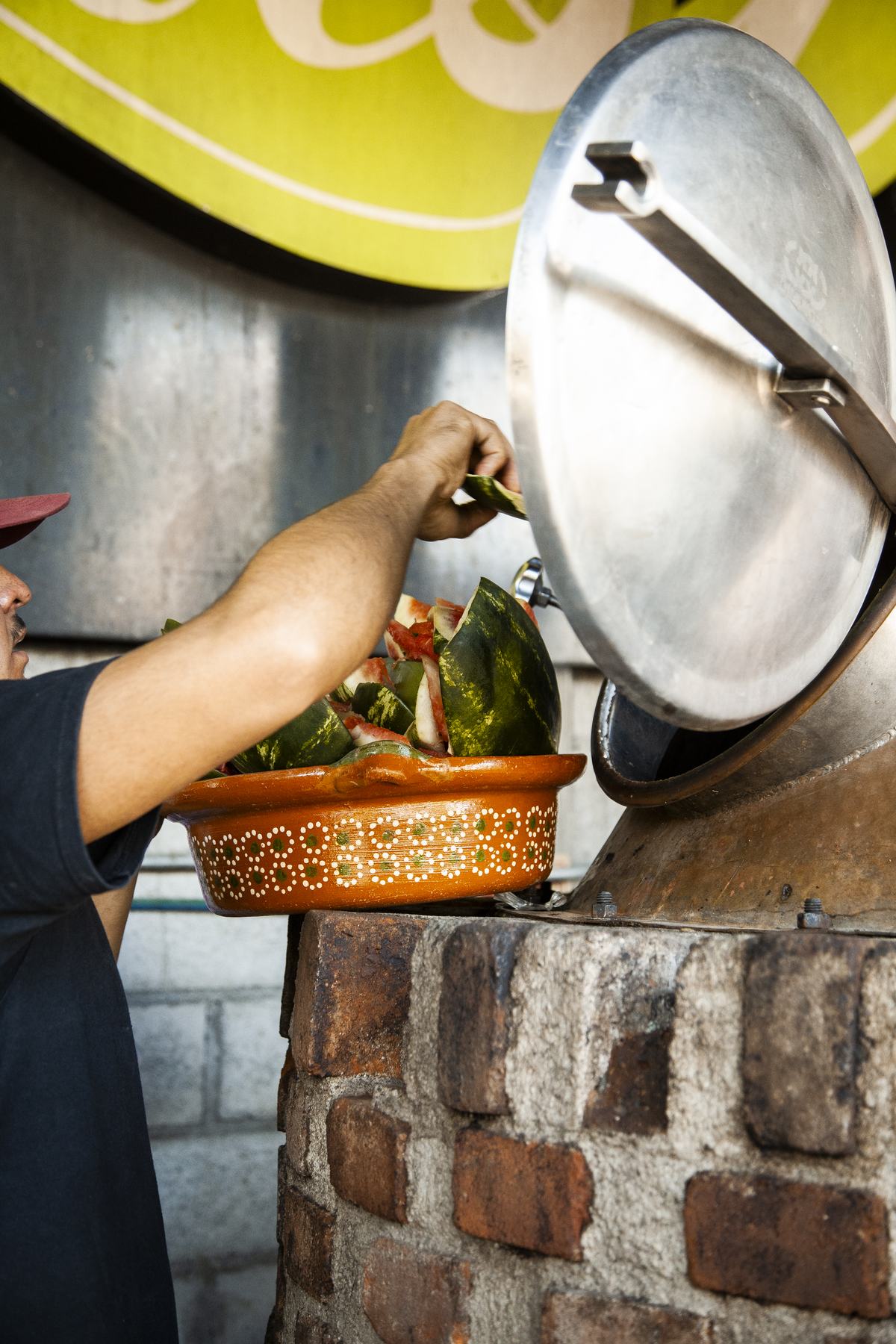 A producer tips a painted clay bowl of cut watermelon into the open hatch of a still.